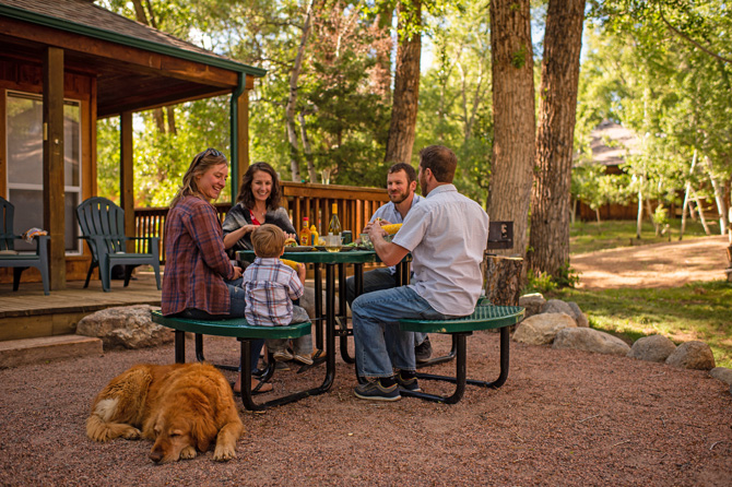 Family with dog eating at a shaded picnic table next to cabin with porch and bbq at Creekside Chalets and Cabins in Salida, Colorado