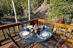 View of private deck with table and chairs along a river and pine tree forest setting at Pinebrook Vacation Rentals in Estes Park, Colorado.
