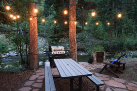 Picnic bench, lounge chair, table and bbq grill lit up at night with a riverfront view at River Bend Mountain Retreat located in Allenspark, Colorado.
