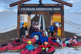 Kids on sledding tubes and adults standing around information sign framing the magic carpet conveyor lift at Colorado Adventure Park near Winter Park in Fraser, Colorado