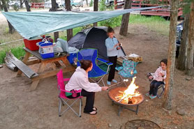 Family making s'mores and relaxing around portable campfire pit at large tent site with picnic table at Surgarbrush Campground in Howard, Colorado.