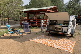 Open teardrop rental camper trailer parked in campsite with lounge chairs, gazebo, picnic tables, with full electric and water hook-ups at Surgarbrush Campground in Howard, Colorado.